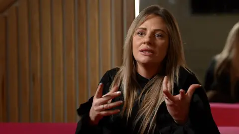 A woman with long, straight, dark blonde hair sits speaking to a camera. She is wearing a long-sleeved black jumper and is gesticulating with her hands. She is sitting on a red sofa, in front of a wall made up of wooden panelling.