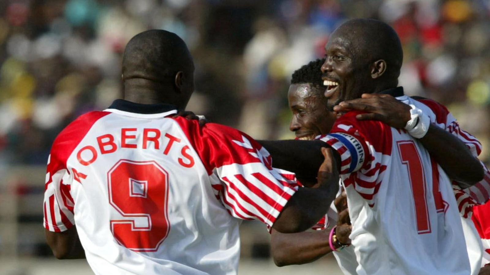George Weah has a broad smile on his face as he celebrates a goal with his Liberia team-mates at the 2002 Africa Cup of Nations. Zizi Roberts, wearing a white shirt with red detail on the sleeve and a red number 9 on the back is seen from behind embracing Weah, while another player who is partially seen has an arm around the striker's shoulders. All three are visible from waist up