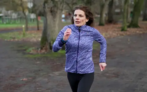 Kristian Johnson/BBC Writer Ruth Clegg, a woman with brown hair in a purple sports jacket runs on a track through a park.