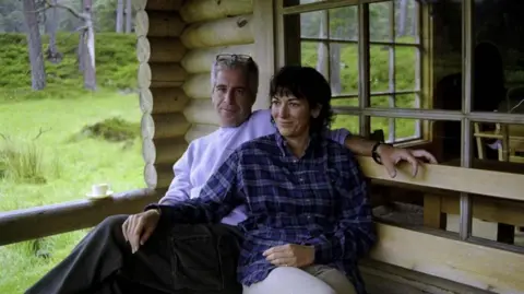 US Attorney's Office SDNY Epstein, a man with grey hair and glasses perched on his head, sitting on a bench outside a log cabin on the Balmoral estate, wearing a pale sweatshirt. His left arm is around Maxwell's shoulder, who rests her hand on his knee. Maxwell has short brown hair and wears and blue checked shirt.