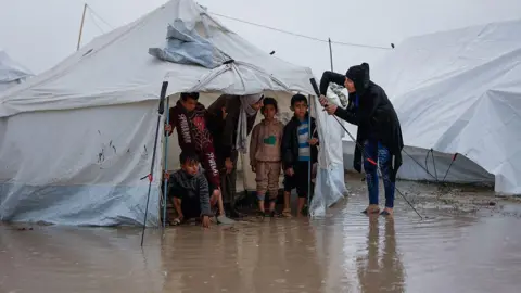 Getty Images A woman fixes her tent as children stand inside at a makeshift camp sheltering displaced Palestinians after heavy rains in the Zeitoun neighbourhood of Gaza City on December 11, 2025