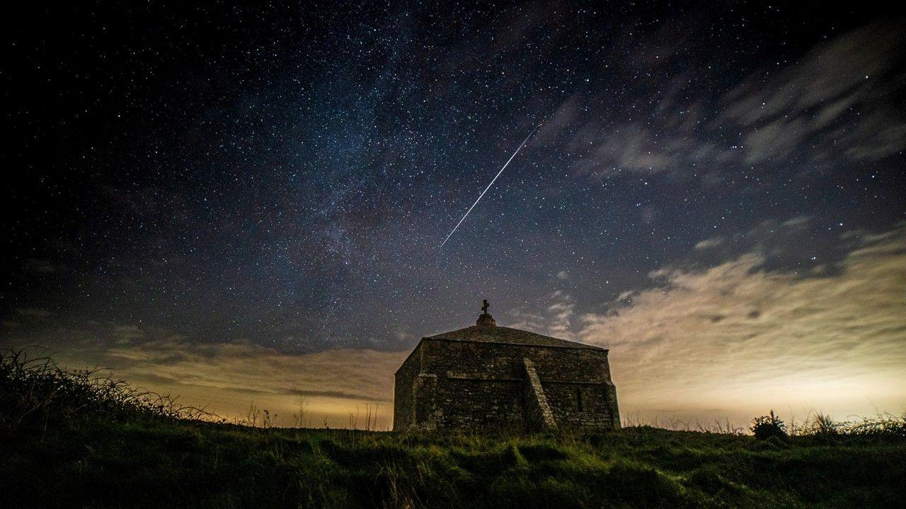 A stone square tower surrounded by grass, amidst a dark blue sky with stars and the white streak of a meteor shower, with white blanket of cloud underneath
