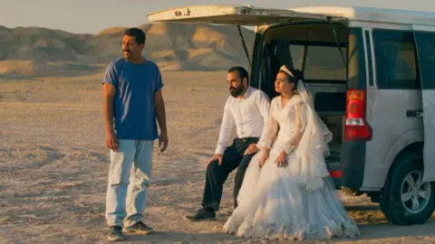 Les Films Pelleas A bride and groom sitting in the back of a car with its boot open, while another man stands beside them, in a promotional image from It Was Just An Accident