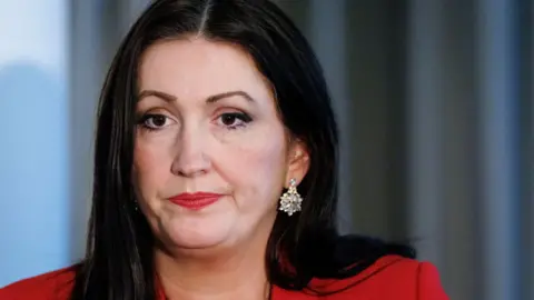 Reuters A close-up of Emma Little-Pengelly sitting at a table. She has long dark hair and has a neutral expression on her face. She is wearing a pair of diamond earrings and a red suit blazer.