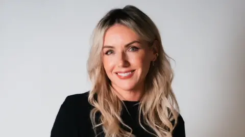 Dora Paphides Sali wears a black top and is smiling at the camera. It's a professional headshot.  She has long wavy blonde hair.