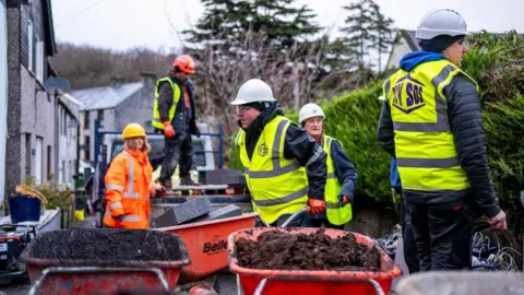 South Shore Construction workers in high-vis jackets marked “DIY SOS” and hard hats can be seen working on a residential street. Several orange wheelbarrows filled with soil and paving blocks are pictured in the foreground. Houses line the street on the left, with a hedge and trees visible on the right.