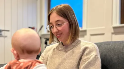 Woman with auburn bob hair smiling at a baby on her knee. She is wearing glasses and a beige wool jumper.