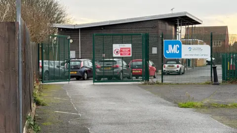 Richard Knights/BBC A general view of the John Mansfield Centre. There are cars in a car park in the foreground, behind a wire fence, and the centre building is in the background. It is a dark brick structure with a white roof.