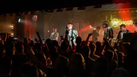 Getty Images Sam Fender and his band performing on a stage at King Street Social Club. He is being cheered on by a crowd, many of whom are holding their arms aloft.
