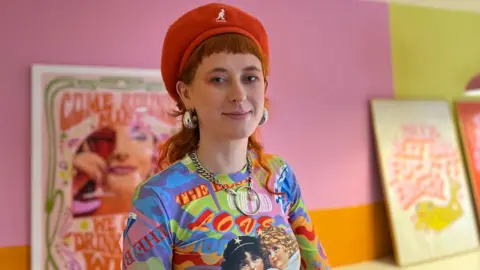 Alice Cullinane/BBC A woman with long red hair is wearing a red beret, large gold earrings and a long sleeved graphic top. She is standing in front of a pink, orange and yellow background.