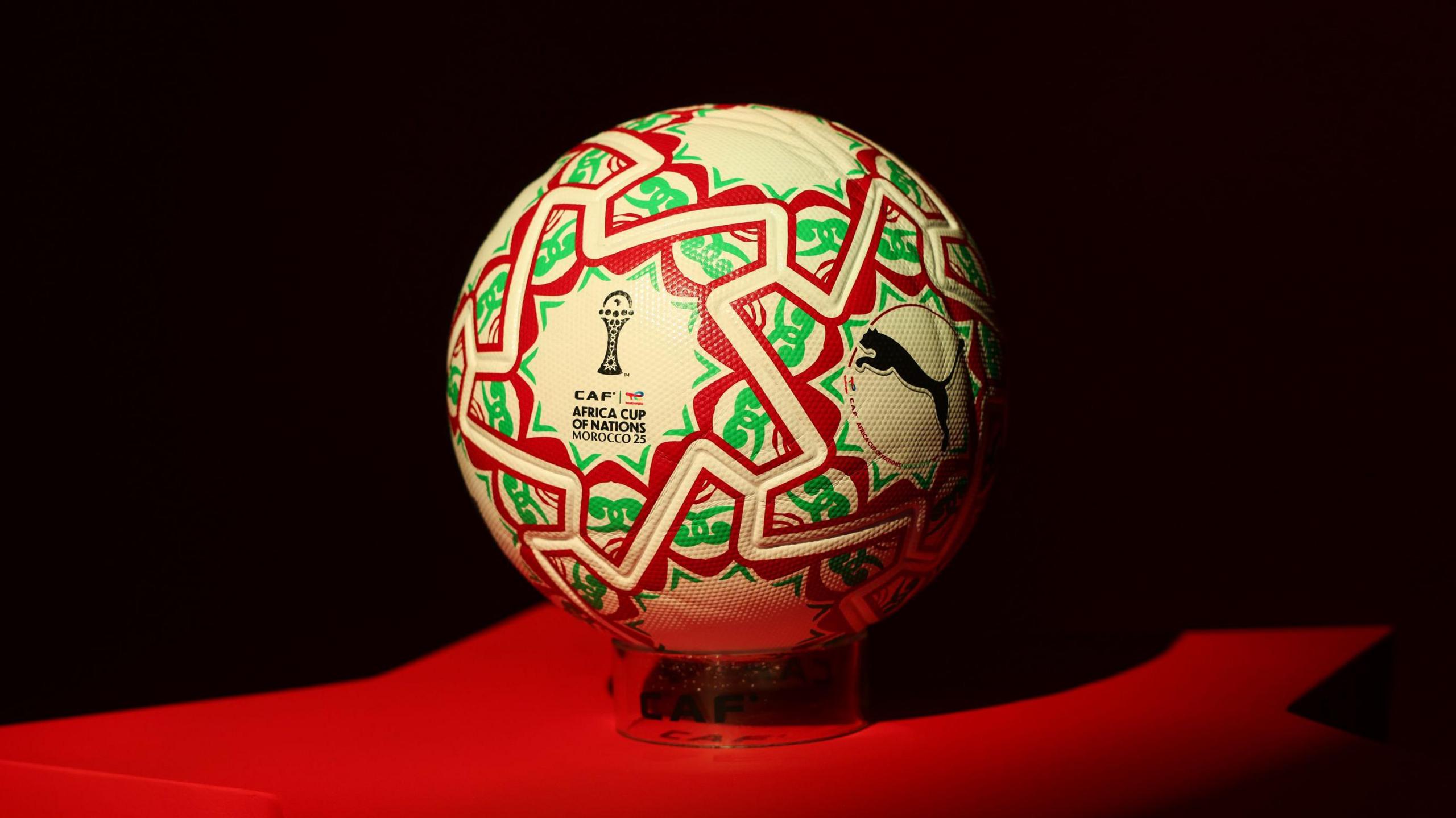 A football stands on a red plinth against a black background. The ball has the Africa Cup of Nations and Puma logos on it, and is decorated with white, green and red patterns