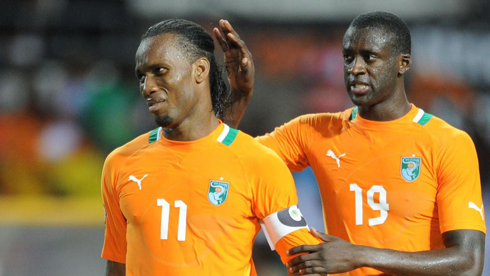 Yaya Toure comforts Didier Drogba during the 2012 Africa Cup of Nations final by going to pat the back of his head with his right hand and holding his left hand on Drogba's left arm. Both players wear bright orange Ivory Coast shirts, and Drogba has a pained expression on his face