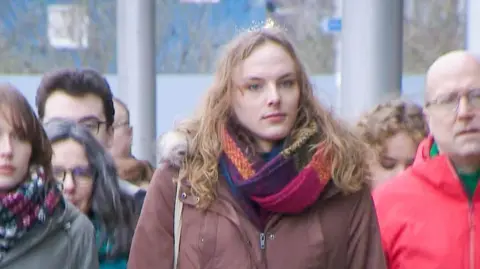 Dr Beth Upton walking outside a court building, dressed in a dark tan coat and wearing a multi coloured scarf. Several supporters are walking alongside.