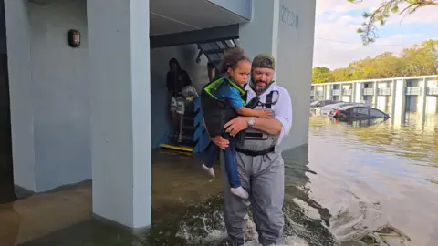 Grey Bull Rescue/Handout Bryan Stern holding a small child in a flooded area in Tampa, Florida during Hurricane Milton in 2024. Water is up to his knees and cars can be seen submerged in the background.
