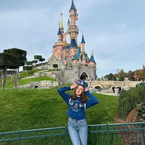 Theme Park Kate A girl wearing Disney ears on her head is posing in front of the Disney castle
