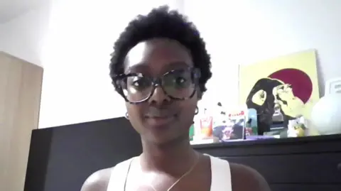 Meerah, a 22-year-old black woman. She is standing in front of a white wall, a television and a fireplace with various art pieces on the mantelpiece