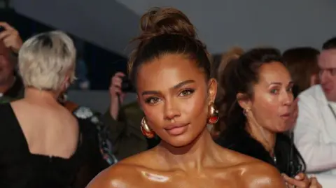 Getty Images Photograph of Tennessee Thresher, who has very large gold earrings on. She is of mixed race ethnicity, has brown hair and a shiny decolletage. She has hazel brown eyes and smiles at the camera. People can be seen in the background behind her.