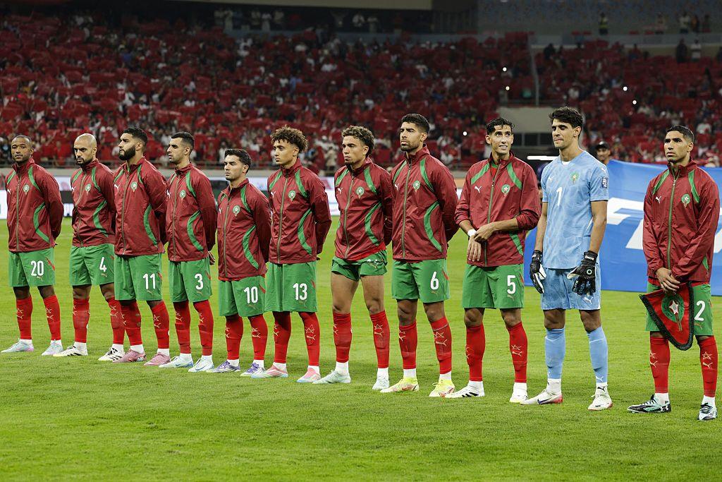 Morocco's men&rsquo;s national football team lined up, during the national anthem, on the pitch before a match. There are 11 players standing shoulder to shoulder in a straight line. Most are wearing Morocco&rsquo;s traditional home colours: red jackets with green detail, green shorts and red socks. The goalkeeper stands out in a light blue kit with gloves on, positioned slightly apart but still aligned with the team. A packed stadium forms the background, with large sections of red-clad supporters filling the stands, reinforcing a home or strongly supported setting