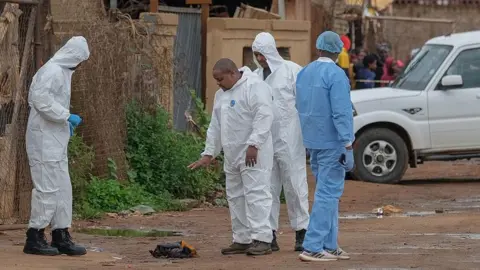 AFP via Getty Images South African Police Service Forensic Pathology Services members in white and blue overalls gesture at the scene of an attack. They are pointing at a pile of clothes on the muddy ground.
