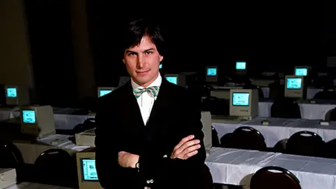 Getty Images A man in a dark suit and green and yellow striped bow tie. He stands in front of a bank of computers with his arms folded.