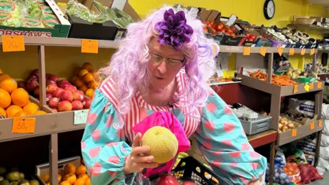 Aunty Pru, aka greengrocer Nick Downes, is in his shop in costume which includes a purple fascinator and a dress with pink polka dots on the sleeves and pink and white stripes on the body. The fruit and veg shop has produce laid out on the shelves behind him and he is holding a melon in his left hand.