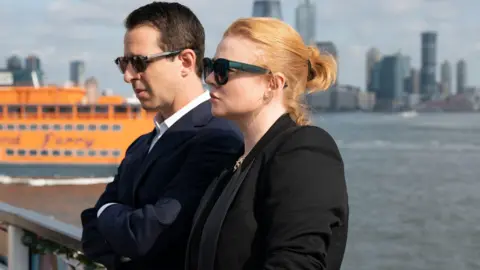 Warner Brothers Discovery Jeremy Strong and Sarah Snook from Succession stand by the water in sunglasses and suits, with New York City in the background.