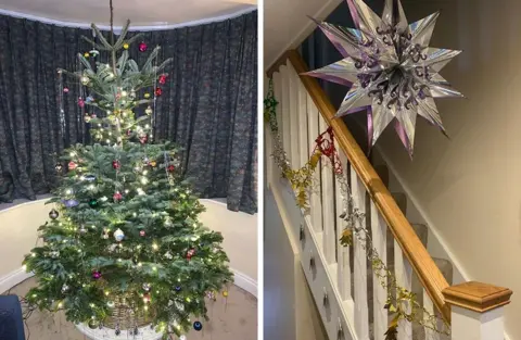 Holly Langley A Christmas tree, indoors, in front of a window with grey curtains. On the tree there are baubles and foil streamers. Stairs with a bannister in a house, with gold foil decorations around the stairs and a silver and purple foil star hanging from the ceiling