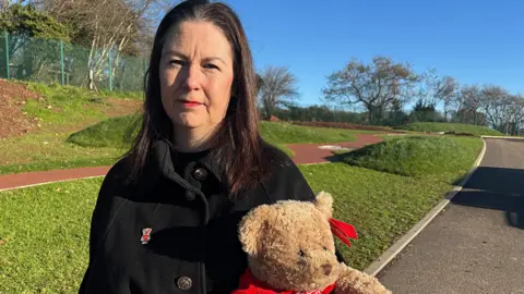 A woman with long dark hair stands in a garden holding a teddy bear. She is wearing a black coat with a bear symbol pinned to it.