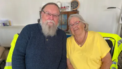 Ross and Anne Campbell sit on their sofa, a cream wall, a display cabinet and a radiator behind them. Ross has thinning hair, a long bears and glasses and Anne has bobbed blonde/grey hair and glasses and wears a bright lemon T-shirt.