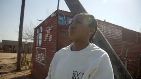 Nthabiseng Nthathakana, in a white jumper, is standing in front of her small shop, which is made of red corrugated iron. She is looking to the left of the picture.
