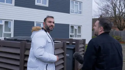 A man in a white coat with a fur hood looks at the camera. He has black-grey hair and a beard. To his right is another man - our BBC reporter - wearing a black jacket and holding up a microphone