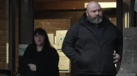 BBC A picture of Hayley Bell, on the left, and Richard Elkin, on the right, coming out of Portsmouth Crown Court. Hayley Bell is wearing a black coat and has long, dark hair. Richard Elkin is bald and has a white and grey beard.