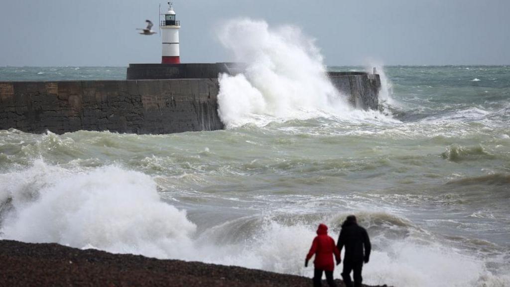 a couple walking along a beach where the sea is looking very choppy with large waves crashing over a sea wall and lighthouse