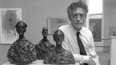 Getty Images Image of Alberto Giacometti in a white shirt and dark tie leaning on a white table with three sculptures of three men. The picture is in black and white.