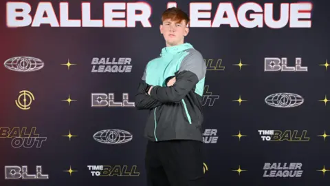 PA Media Angry Ginge wears a blue and grey jacket and black trousers. He has ginger hair and is standing in front of a board that says Baller League