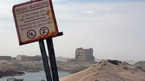 A rusting sign at an angle can be seen in the foreground. In English and Afrikaans it says "Warning, no unauthorised entry, trespassers will be prosecuted." In the background there is an abandoned multi-storey building.
