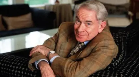 BBC Stanley Baxter is photographed in later life.  He is looking at the camera wearing a suit and he has his arms crossed.