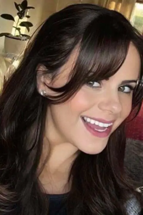 Antonia Merola Jones A woman with long brown hair and wearing a dark top is smiling. A plant, window and cream-coloured curtains can be seen behind her.