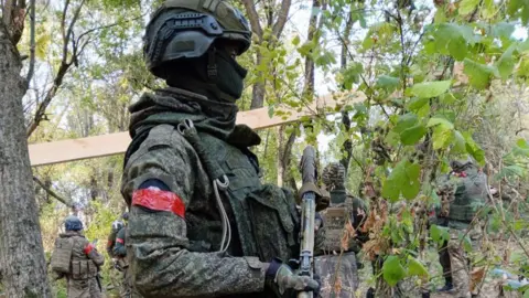 Kuloba family David Kuloba in full battle gear holding a gun in a forest in Ukraine with other soldiers seen in the background.