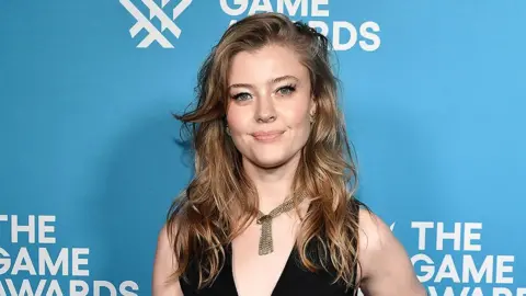 Getty Images A woman in a halterneck black dress stands in front of a light blue backdrop with a "The Game Awards" logo printed on it. Her shoulder-length blonde hair is styled in waves and she wears a flat gold necklace to complete the outfit