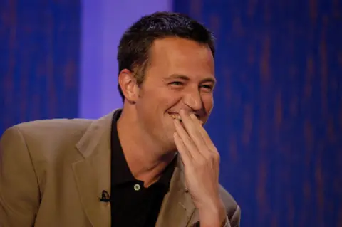 Matthew Perry covers his mouth as he laughs. He wears a tan blazer and black polo shirt