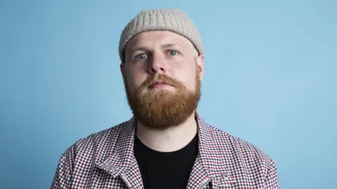A mid-shot of Tom Walker looking into the camera. He is wearing a beige wolly hat and a chequered shirt.