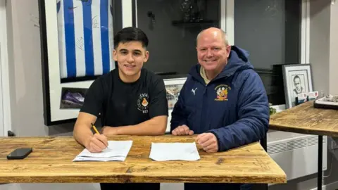 Handout Bryan wears a black t-shirt with a crest and has black hair. He is signing a piece of paper on a wooden table. He is stood beside a man who wears a navy Wythenshawe jacket