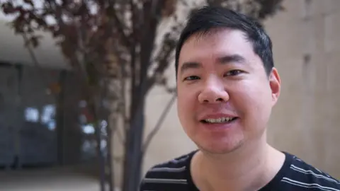 A man with short dark hair standing in front of a building and a tree smiles at the camera.