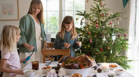 Getty Images A mother and her two daughters set the Christmas dinner table. There is a Christmas tree in their kitchen