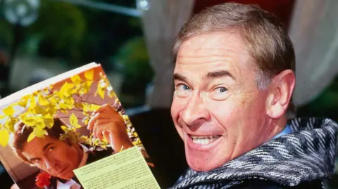 Mirrorpix via Getty Images A man smiles at the camera, looking back over his shoulder while holding a book with his photo on the cover