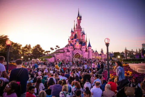 NurPhoto via Getty Images A view of the Sleeping Beauty Castle during the sunset at Disneyland Paris, in Paris, France - crowds gather in front of the castle and the sky and castle are tainted pink.