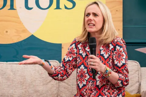 Lorne Thomson/Redferns Mariana Hyde in a flowery red dress holding a microphone while making an expressive hand movement.