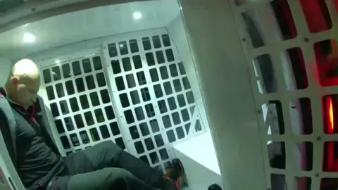 Dyfed-Powys Police Police body camera footage of a man with his arms behind his back in a police vehicle. There's a white grille behind him and the back doors are open. he is dressed all in black. He has his head looking down at his feet. he is bald and clean shaven.