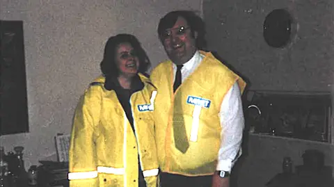 Raynet An old snap of Anne and Ross in their youth, both with dark hair and smiling. They wear yellow high visibility Raynet jackets. It's a blurry old image with a very dark background.